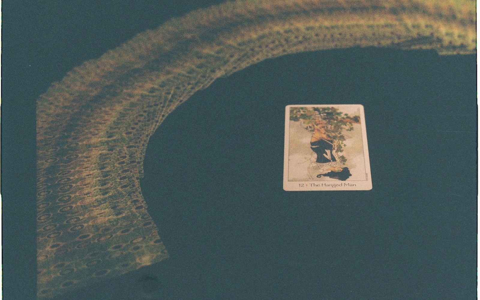 The Hanged Man tarot card rests on a dark surface beside a curved lace pattern