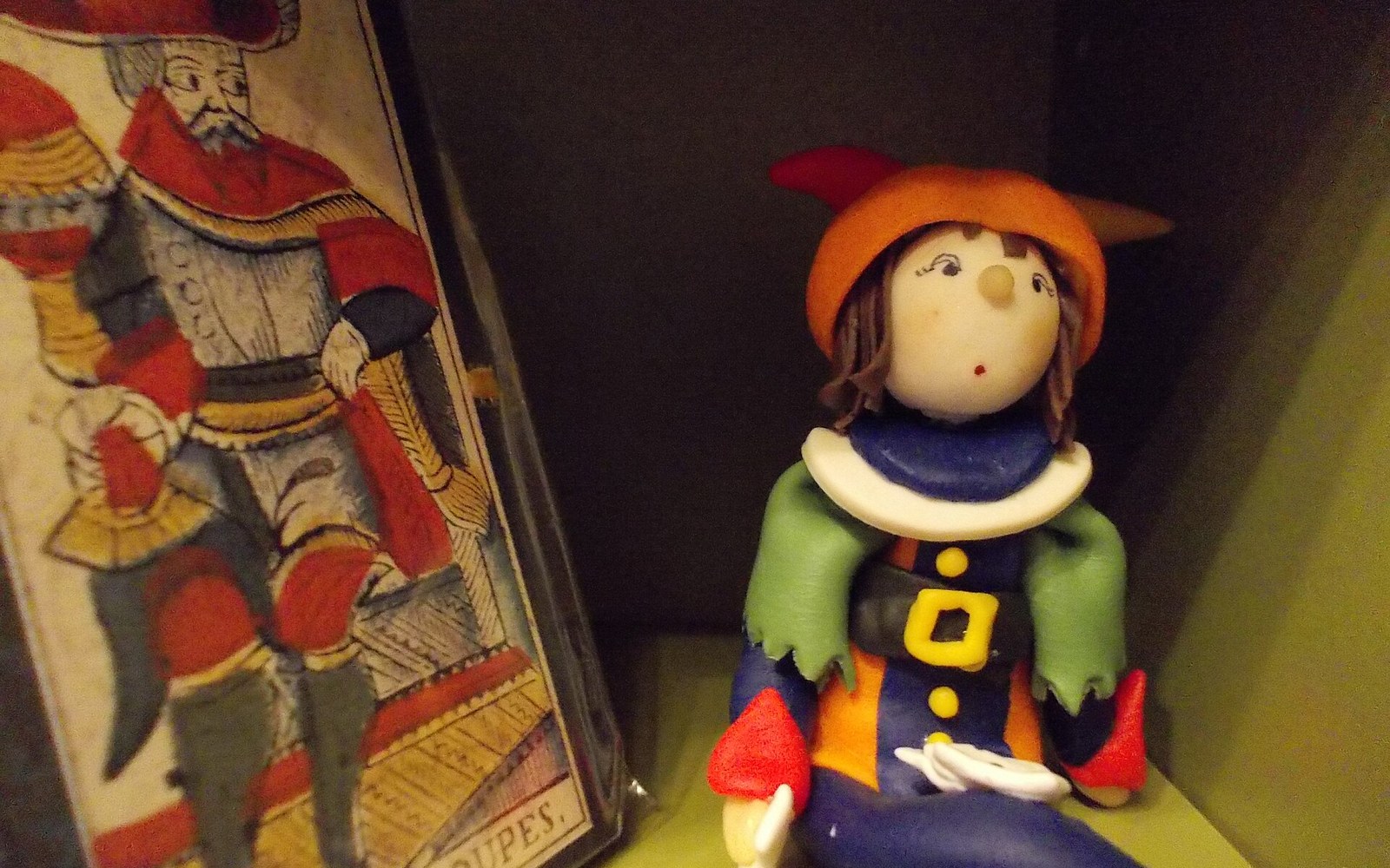 Small jester figurine with an orange hat stands beside an illustrated tarot card